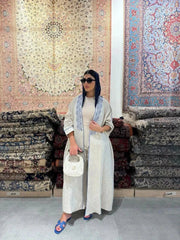 Woman wearing a beige bisht with blue embroidery and piping, styled with a navy headscarf, white handbag, and blue sandals, standing outdoors on a paved street.