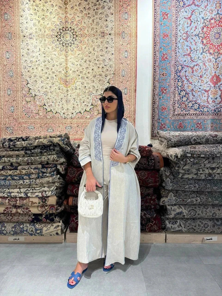 Woman wearing a beige bisht with blue embroidery and piping, styled with a navy headscarf, white handbag, and blue sandals, standing outdoors on a paved street.