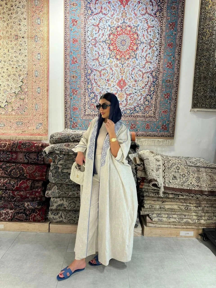 Woman wearing a beige bisht with blue embroidery and piping, styled with a navy headscarf, white handbag, and blue sandals, standing outdoors on a paved street.