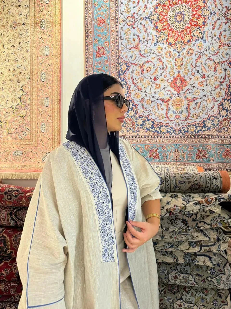 Woman wearing a beige bisht with blue embroidery and piping, styled with a navy headscarf, white handbag, and blue sandals, standing outdoors on a paved street.