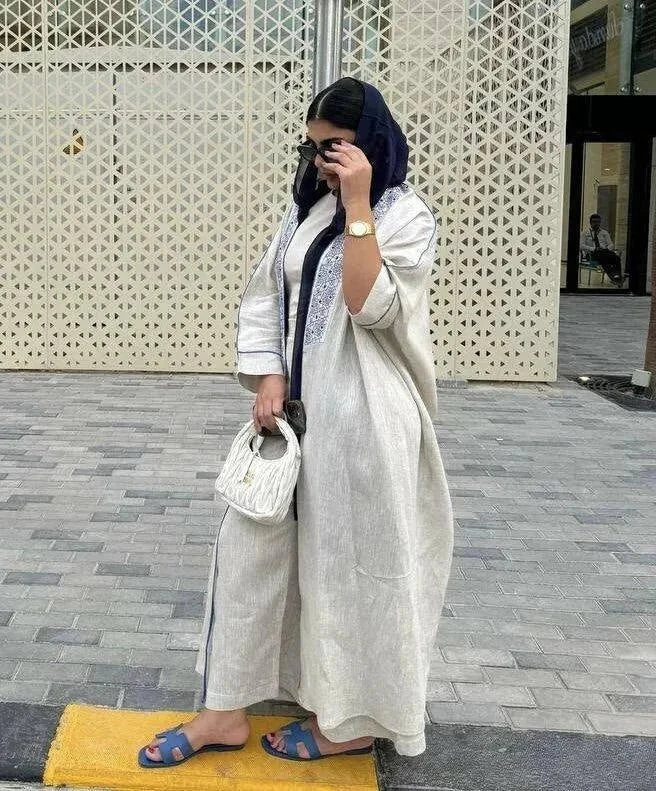 Woman wearing elegant beige bisht with blue embroidery and piping, paired with navy headscarf and casual sandals, standing outdoors on paved ground.