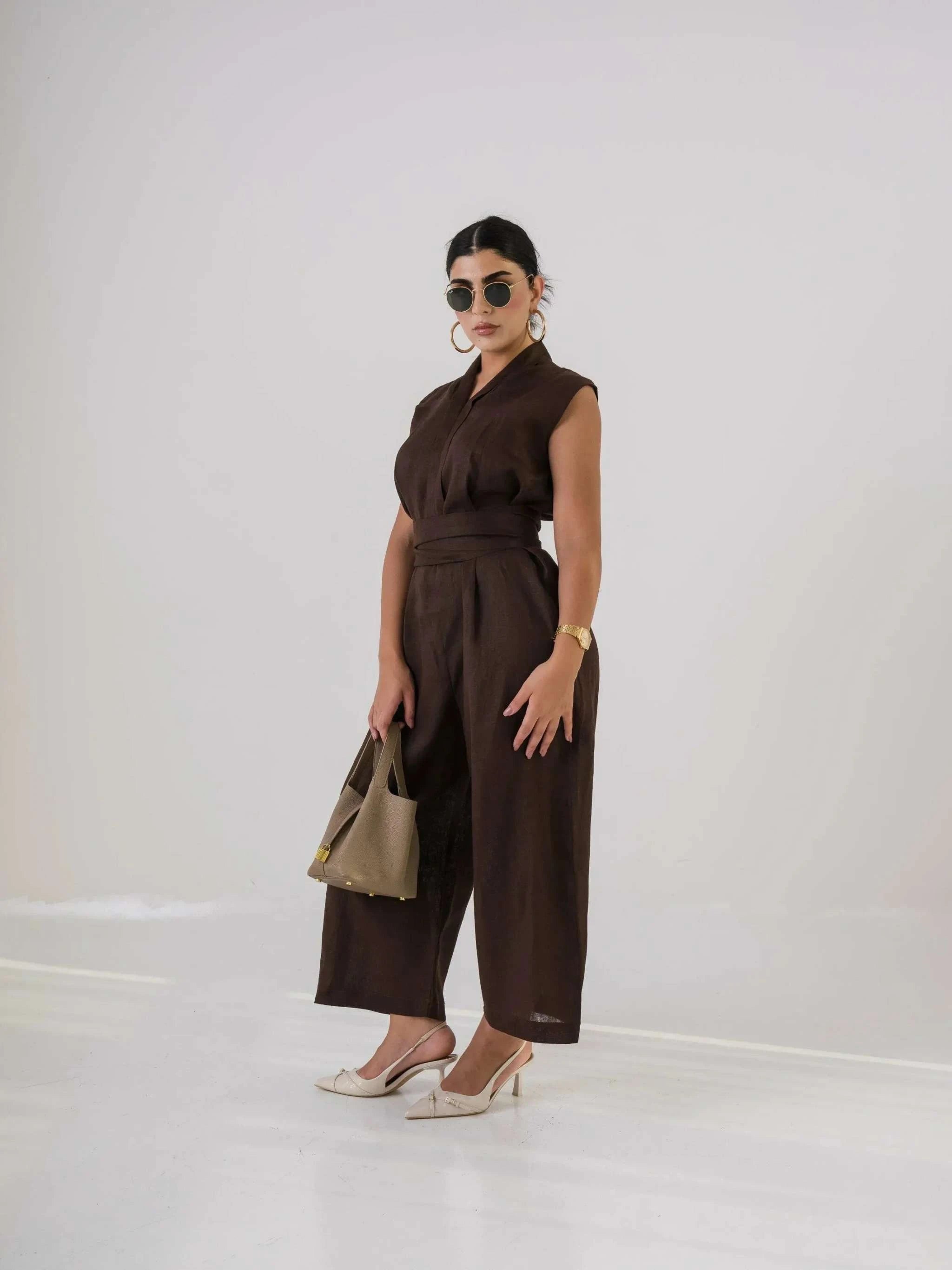 Woman wearing dark brown linen jumpsuit with sleeveless design, holding beige handbag and wearing sunglasses and heels
