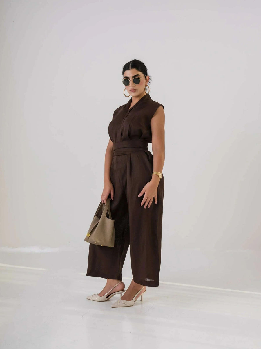 Woman wearing dark brown linen jumpsuit with sleeveless design, holding beige handbag and wearing sunglasses and heels