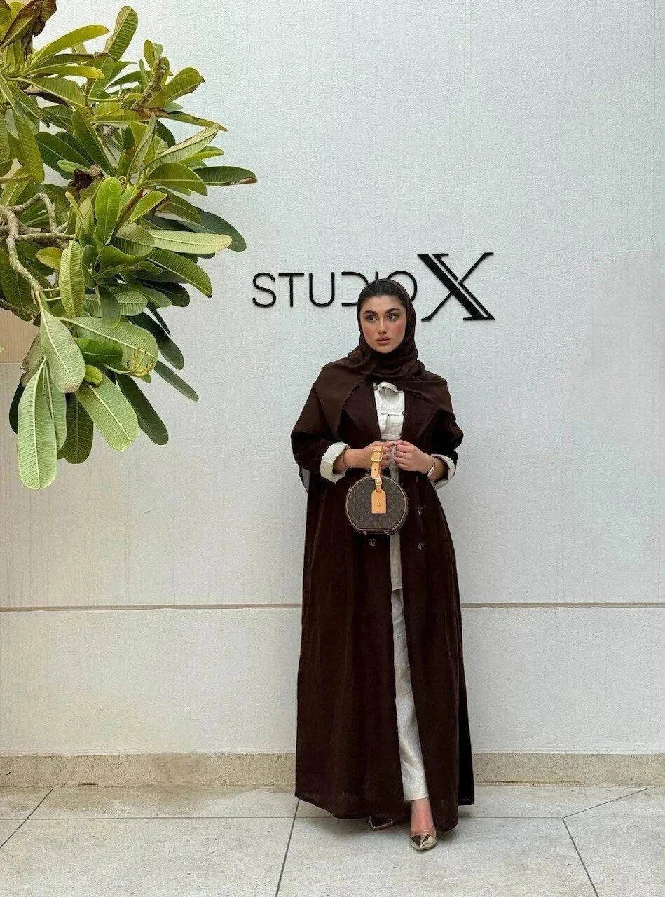 woman wearing a dark brown tailored linen blazer styled as a long coat with a matching headscarf and white outfit underneath standing against a white wall with a green plant nearby - dark brown blazer