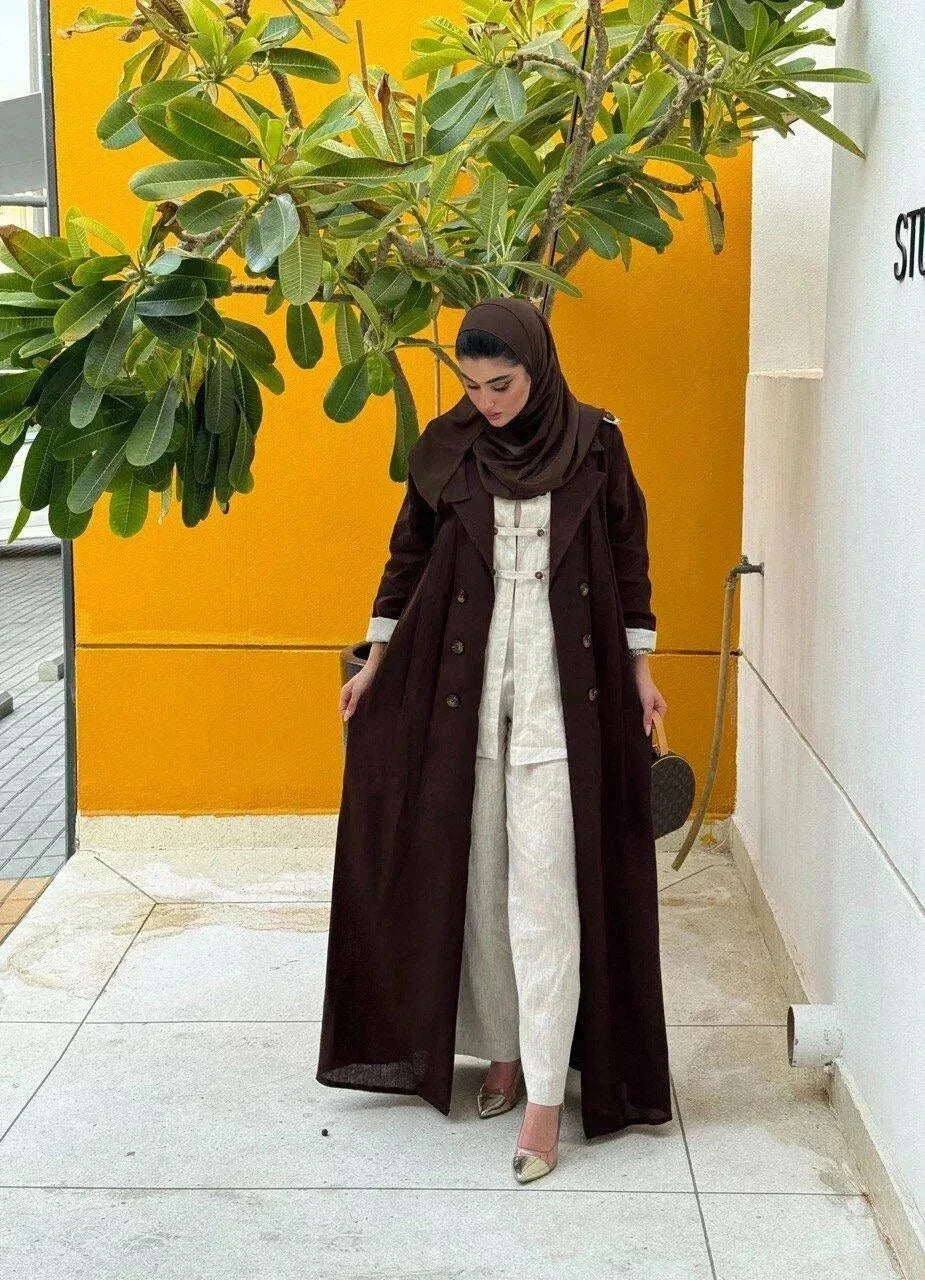 woman wearing a dark brown tailored linen blazer styled as a long coat with a matching headscarf and white outfit underneath standing against a white wall with a green plant nearby - dark brown blazer