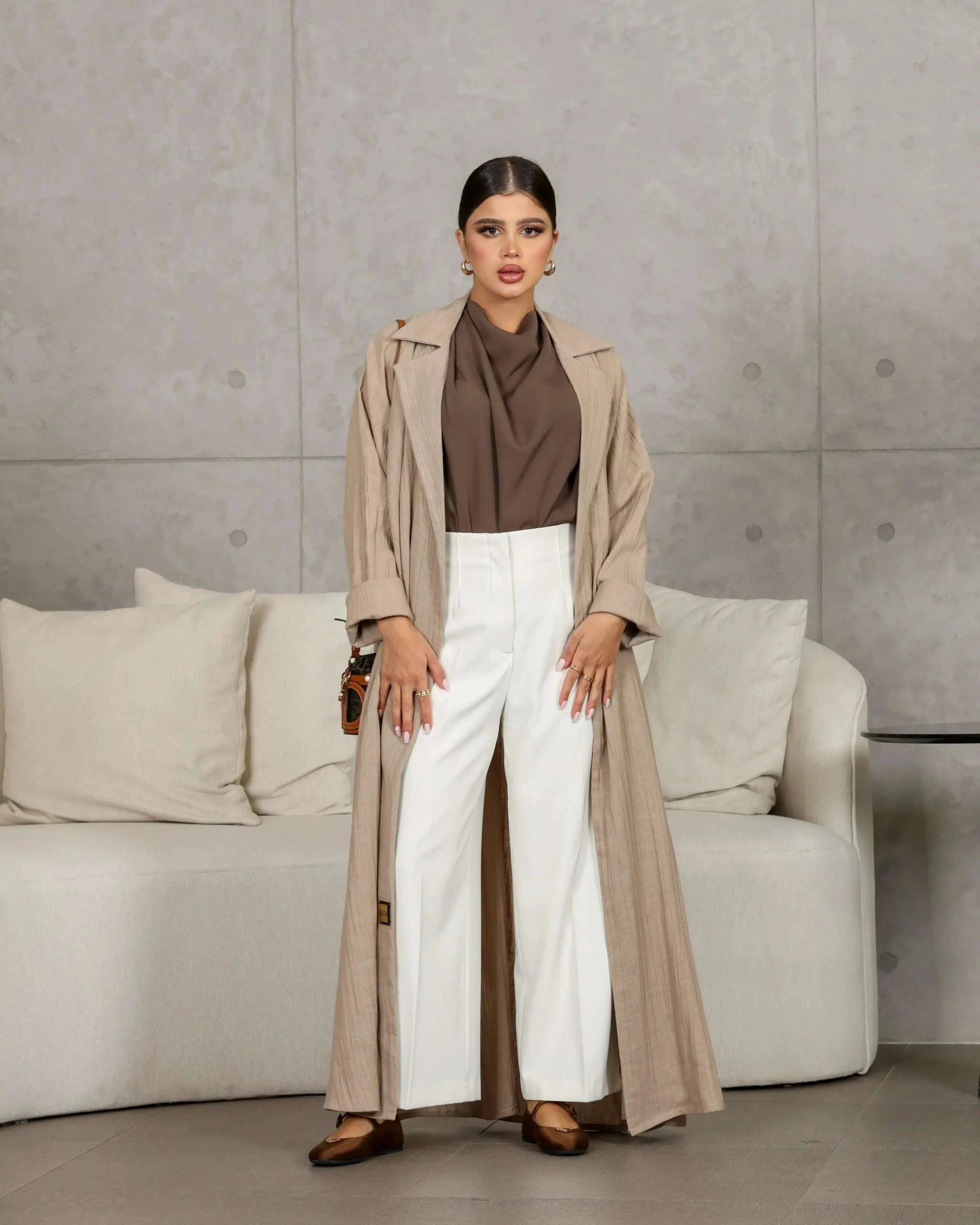 woman wearing taupe relaxed beige abaya made from crumpled linen with white pants and brown top standing indoors