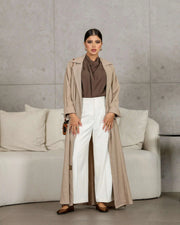 woman wearing taupe relaxed beige abaya made from crumpled linen with white pants and brown top standing indoors