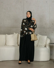 Elegant black abaya with white and earthy orange floral embroidery on bodice and sleeves, worn with a beige handbag in a modern setting