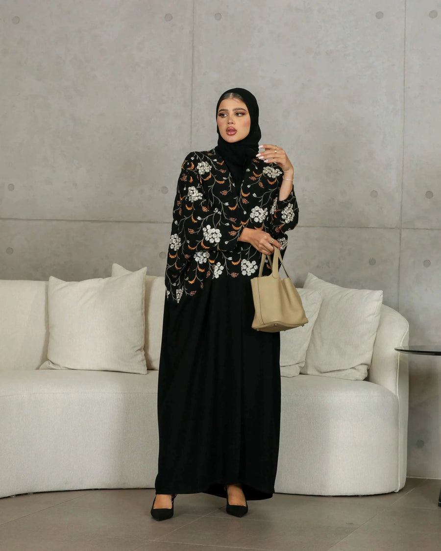 Elegant black abaya with white and earthy orange floral embroidery on bodice and sleeves, worn with a beige handbag in a modern setting