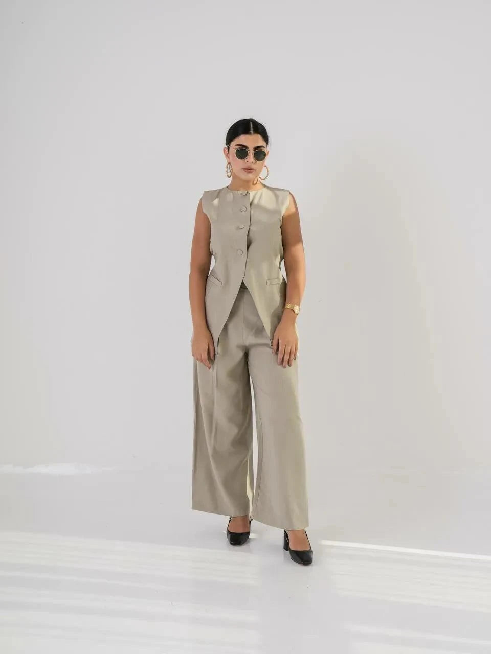 Woman wearing linen beige pants with a matching sleeveless vest and sunglasses in a minimalist setting