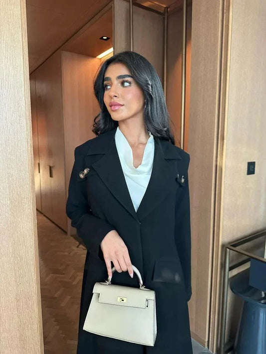 woman wearing black blazer-style abaya with button detailing on shoulders and back holding beige handbag
