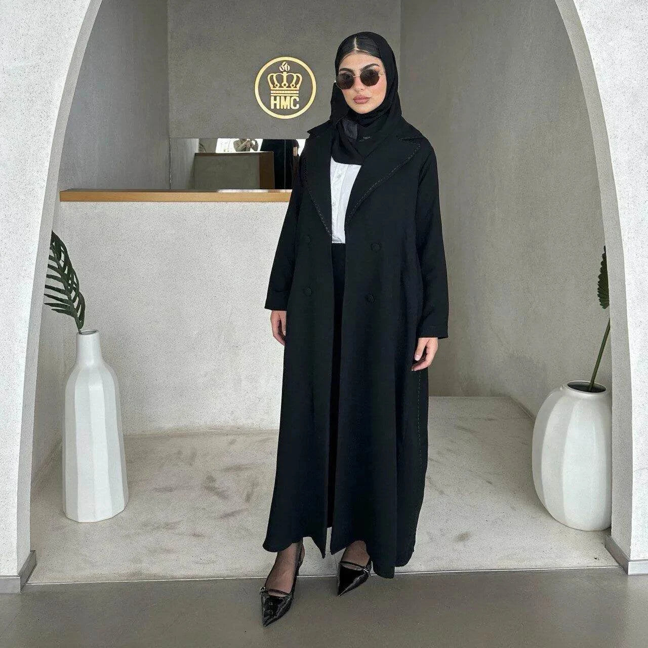 Woman wearing sleek black abaya with subtle black line detailing and hijab in modern minimalist setting