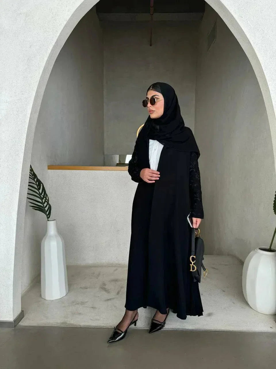 Woman wearing black crepe abaya with embroidered sleeves and pleated detailing, styled with black hijab and sunglasses in modern minimalist setting