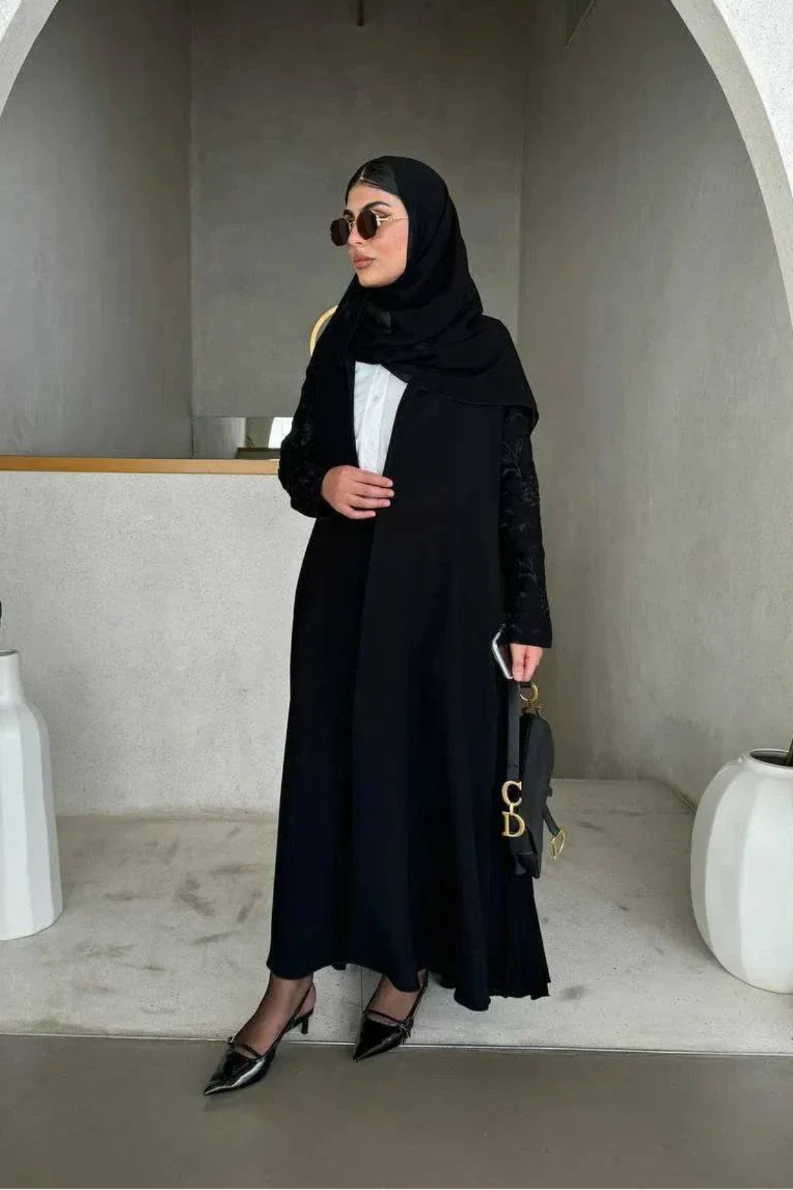 Woman wearing black crepe abaya with embroidered sleeves and pleated detailing, styled with black hijab and sunglasses in modern minimalist setting