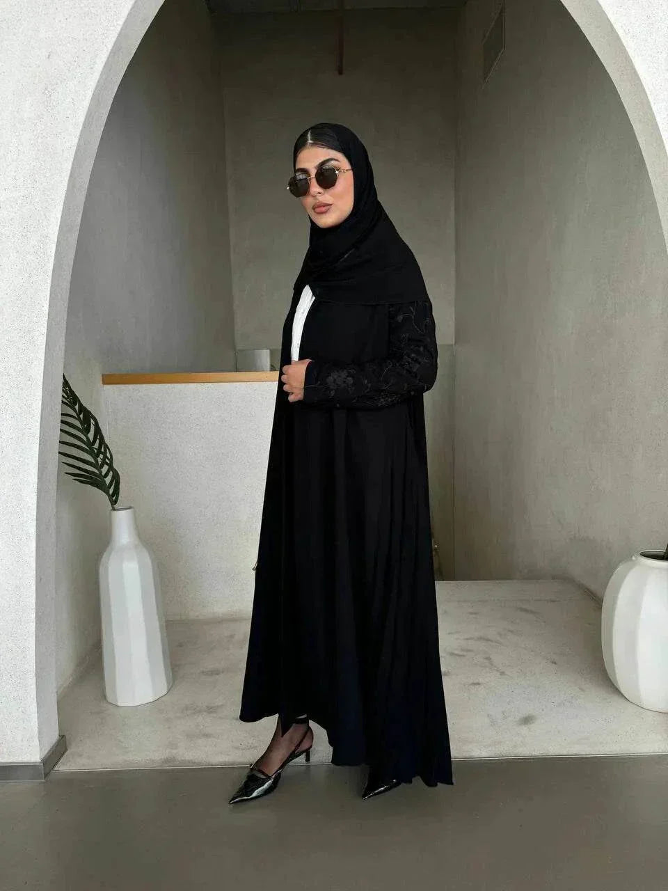 Woman wearing black crepe abaya with embroidered sleeves and pleated detailing, styled with black hijab and sunglasses in modern minimalist setting