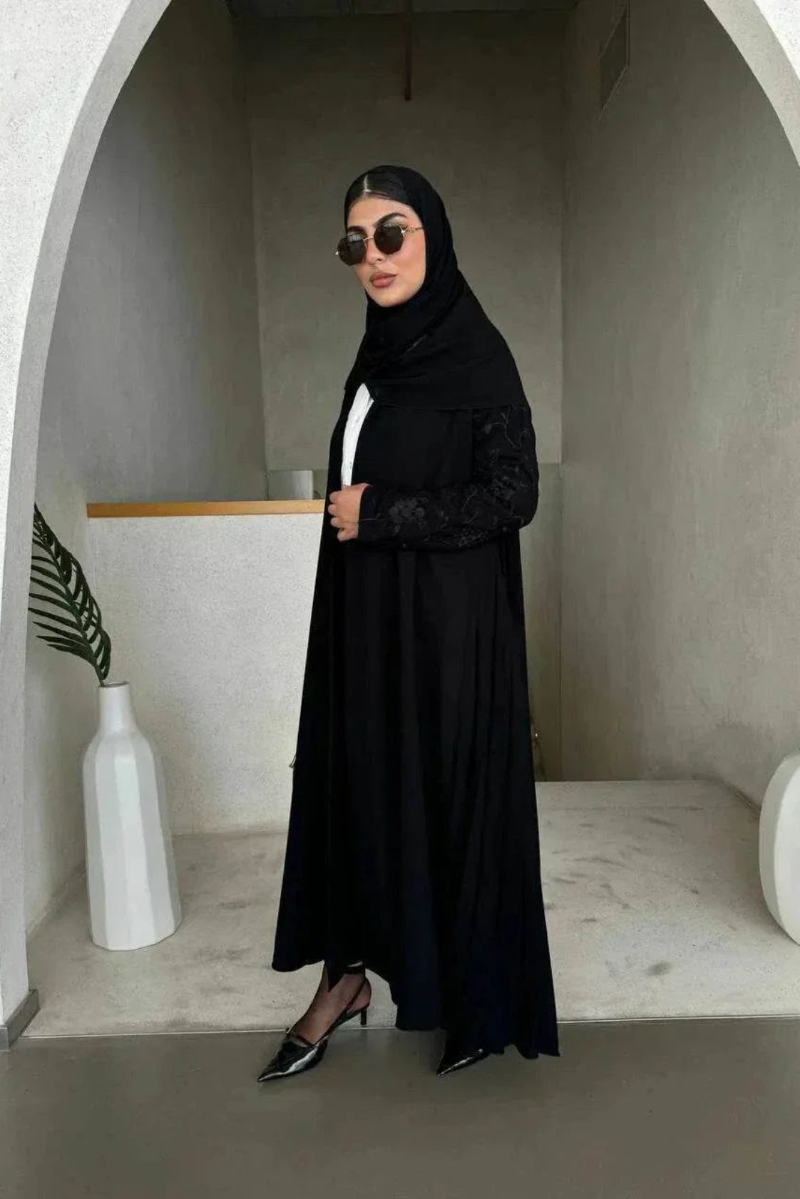 Woman wearing black crepe abaya with embroidered sleeves and pleated detailing, styled with black hijab and sunglasses in modern minimalist setting