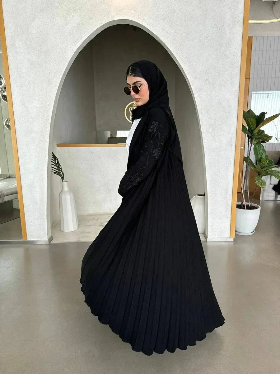 Woman wearing black crepe abaya with embroidered sleeves and pleated detailing, styled with black hijab and sunglasses in modern minimalist setting