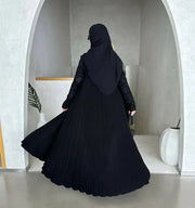 Woman wearing black crepe abaya with embroidered sleeves and pleated detailing, styled with black hijab and sunglasses in modern minimalist setting
