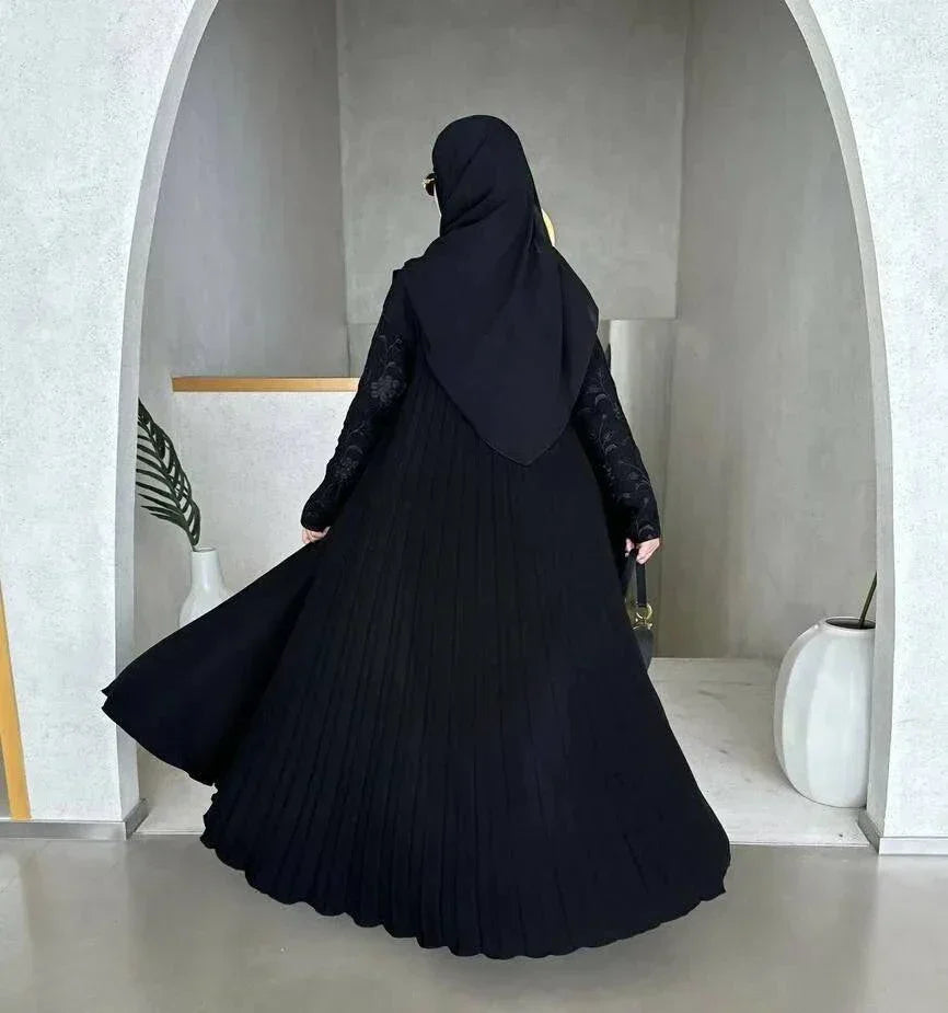 Woman wearing black crepe abaya with embroidered sleeves and pleated detailing, styled with black hijab and sunglasses in modern minimalist setting