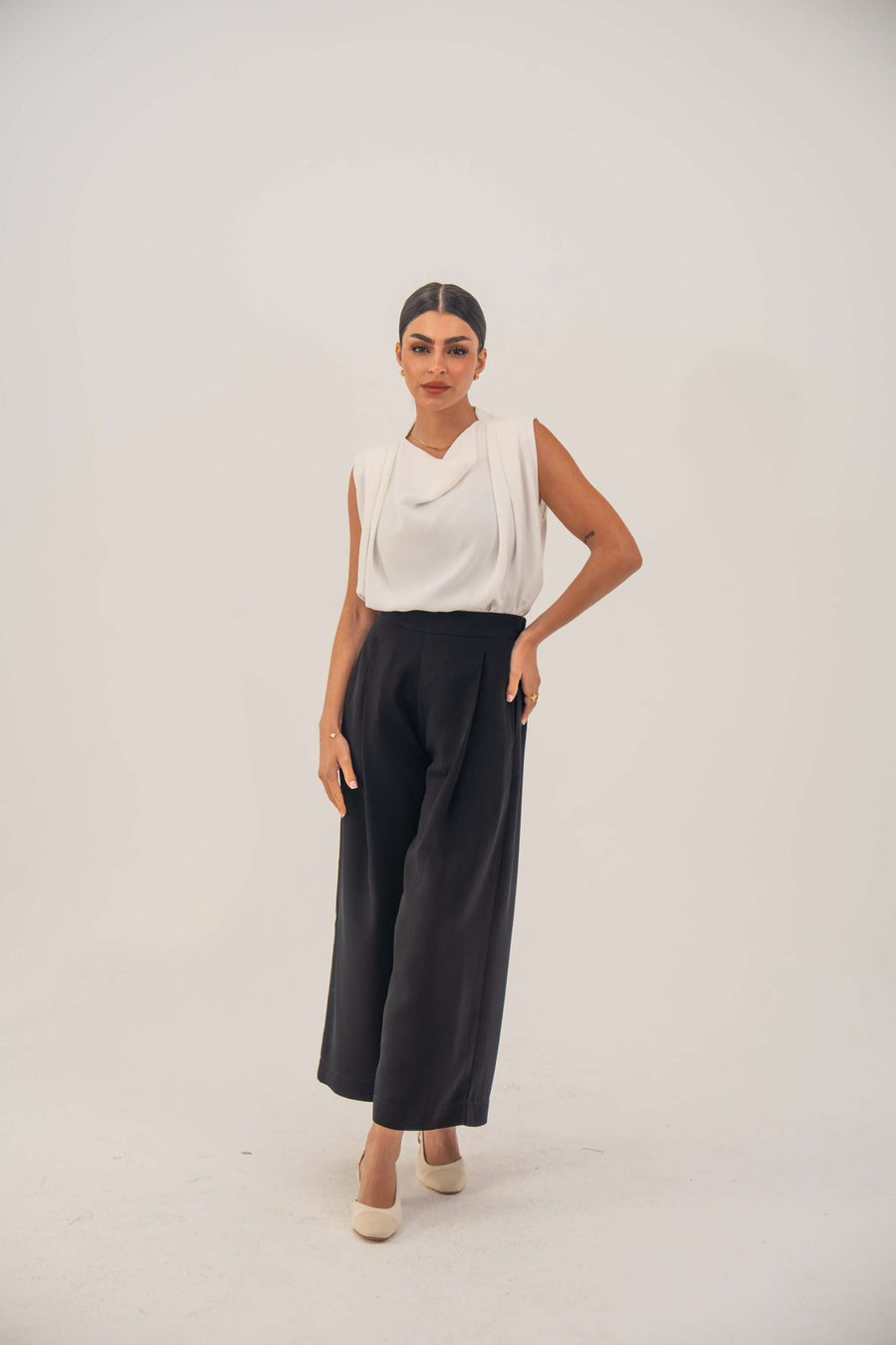 Woman wearing a sleeveless white crepe blouse with a draped neckline and black high-waisted pants