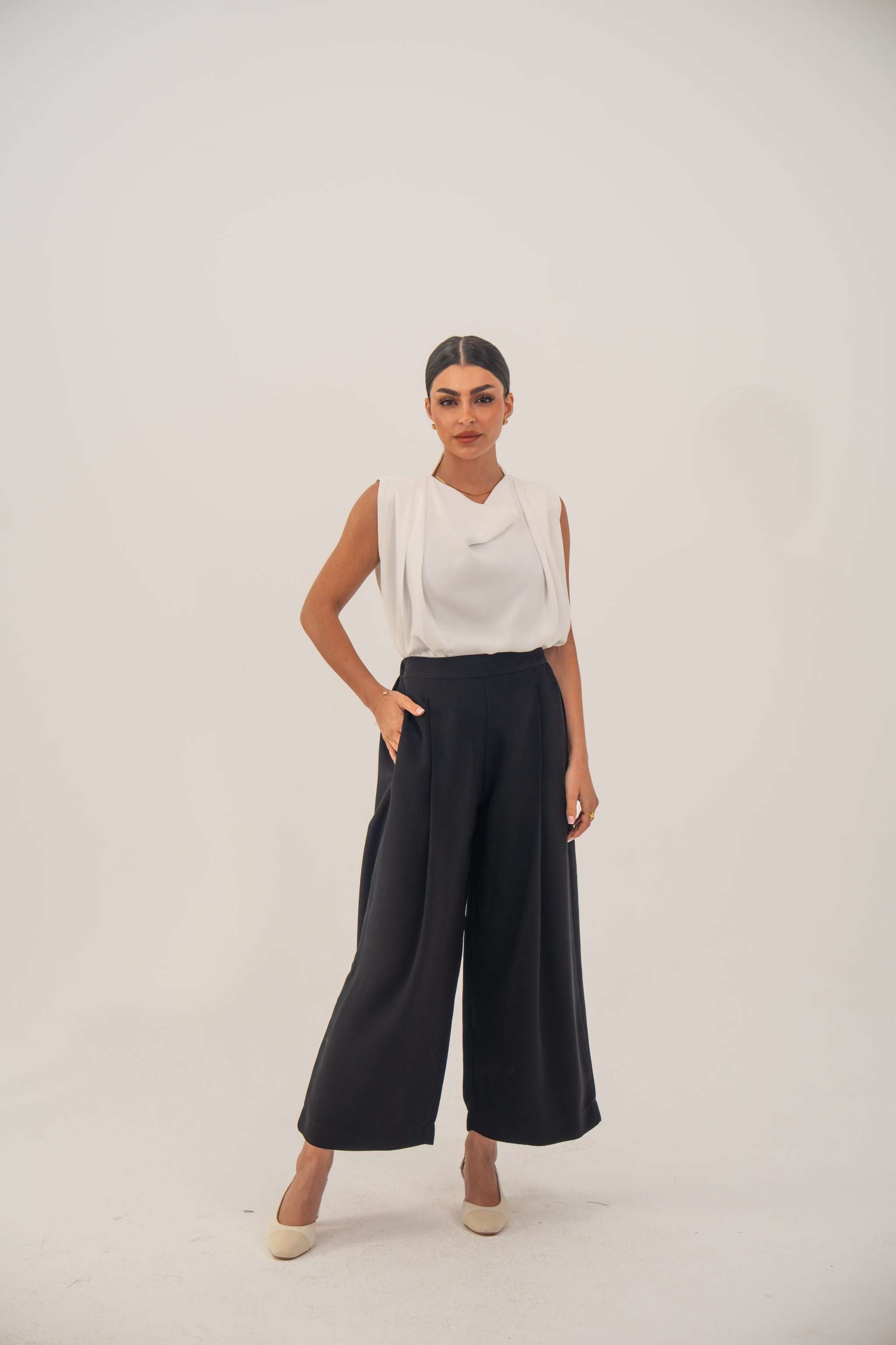 Woman wearing a sleeveless white crepe blouse with a draped neckline and black high-waisted pants