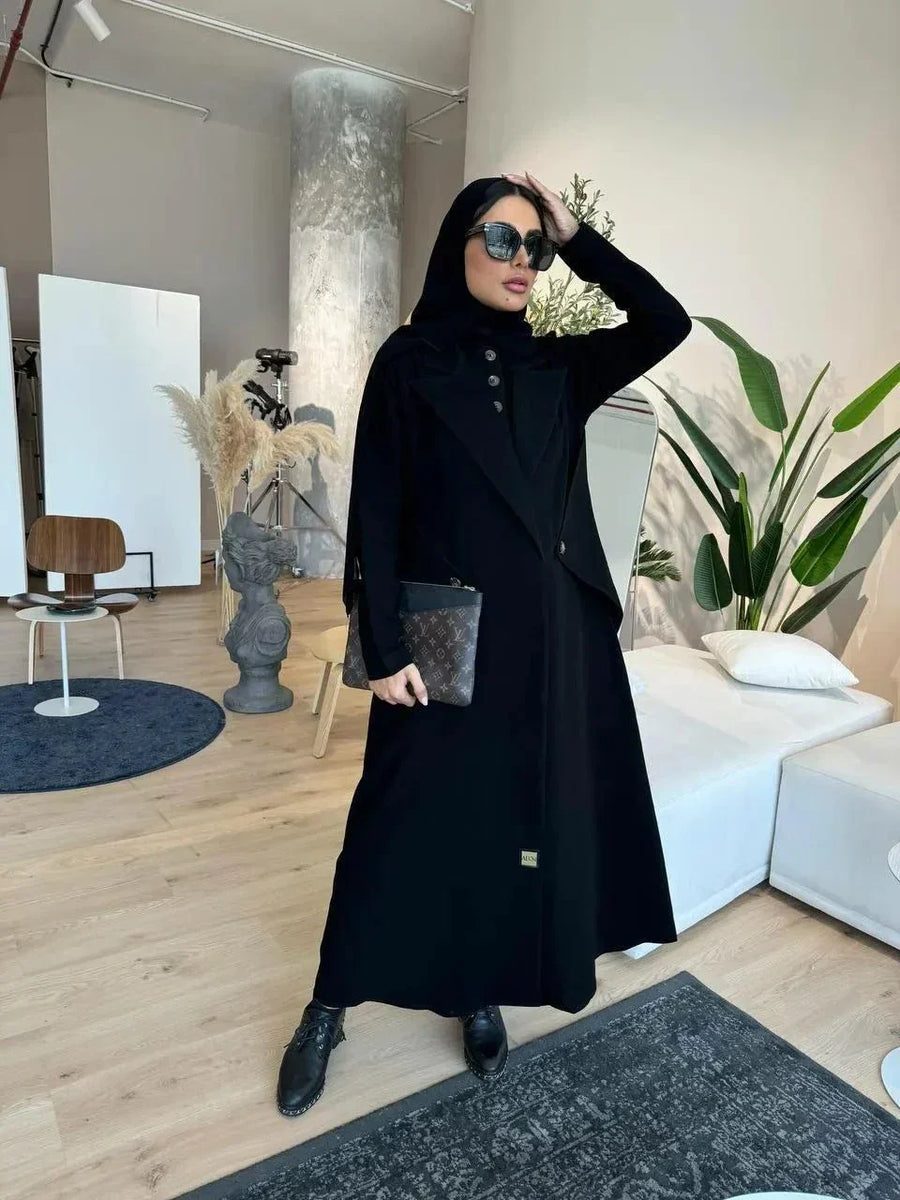 Black abaya with blazer-inspired tailoring, sharp lapels, buttoned front, and angled side flap pockets in a modern indoor setting.