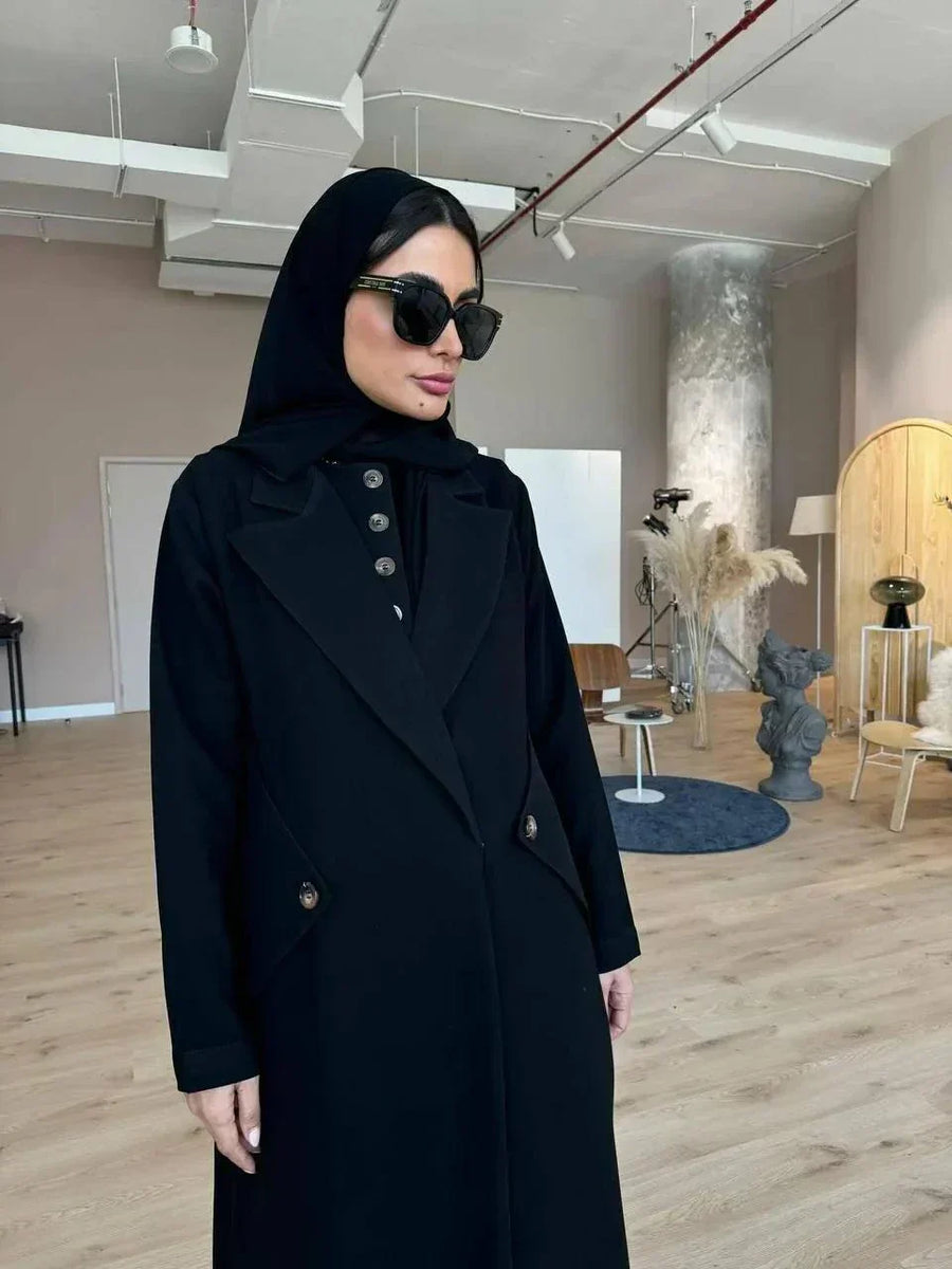 Black abaya with blazer-inspired tailoring, sharp lapels, buttoned front, and angled side flap pockets in a modern indoor setting.
