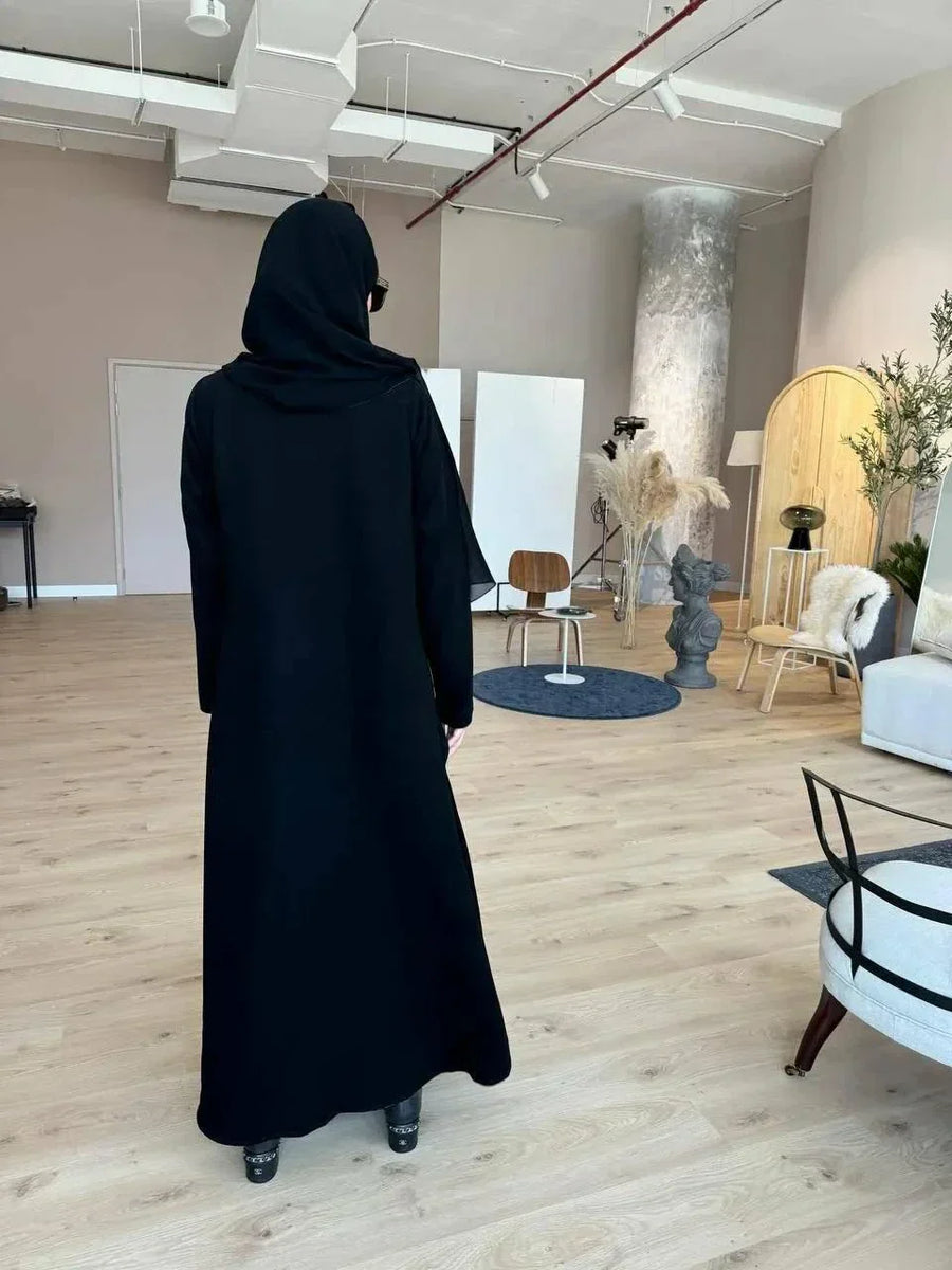 Black abaya with blazer-inspired tailoring, sharp lapels, buttoned front, and angled side flap pockets in a modern indoor setting.