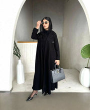 Woman wearing Obscia black abaya with blazer-style trench collar and structured shoulder flaps, holding a gray handbag and sunglasses.