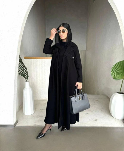 Woman wearing Obscia black abaya with blazer-style trench collar and structured shoulder flaps, holding a gray handbag and sunglasses.