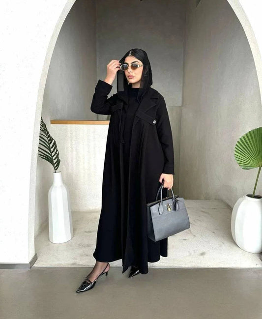 Woman wearing Obscia black abaya with blazer-style trench collar and structured shoulder flaps, holding a gray handbag and sunglasses.
