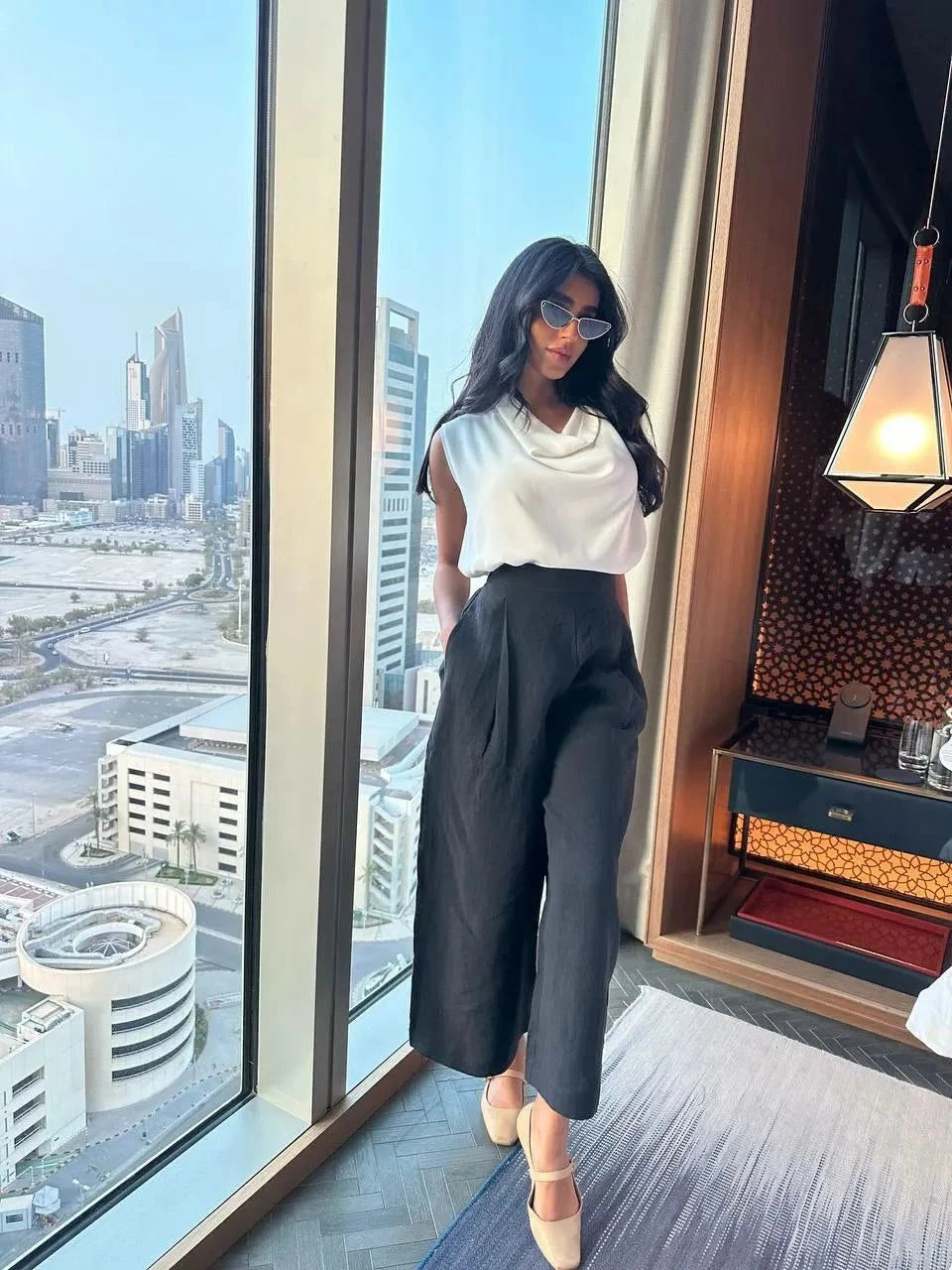 Woman wearing relaxed fit washed linen black pants with a white sleeveless top standing by a large window overlooking a cityscape