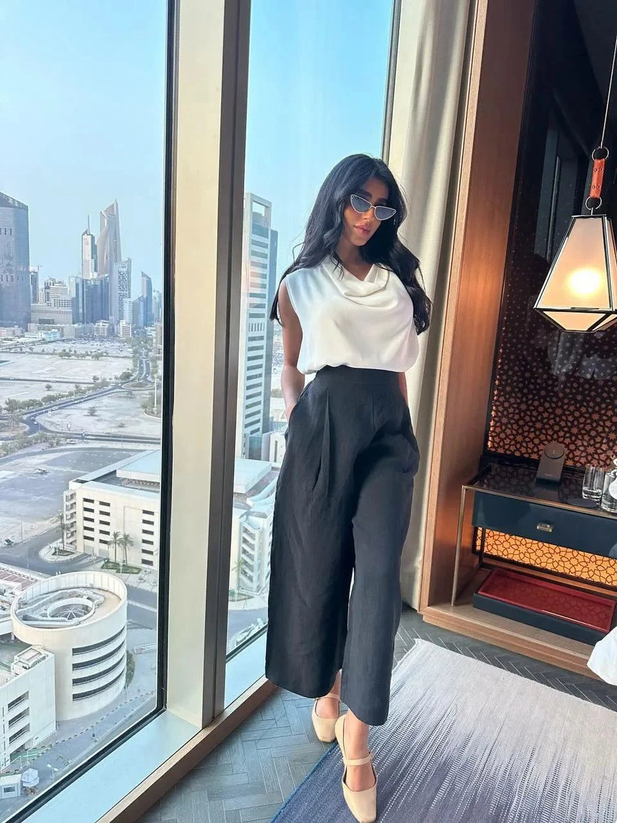 Woman wearing relaxed fit washed linen black pants with a white sleeveless top standing by a large window overlooking a cityscape