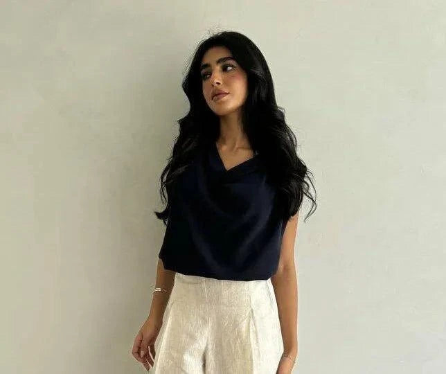 Woman wearing a smooth black crepe blouse with a draped neckline and light-colored pants against a plain background.