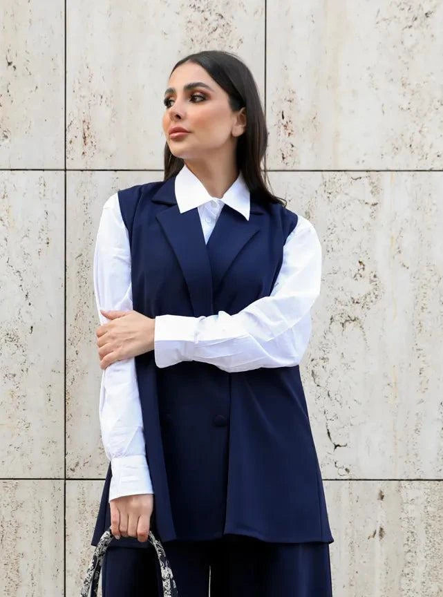 Woman wearing navy vest and wide-leg pants set with white shirt standing against a beige stone wall