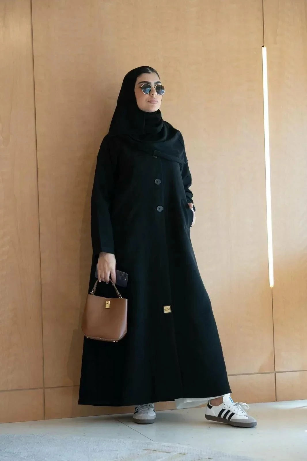 Woman wearing a black crepe abaya with three front buttons, paired with sunglasses and a brown handbag.
