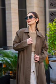 Woman wearing a long coffee-cWoman wearing a long coffee chino coat with tailored fit and sunglasses in an outdoor urban settingolored chino coat with tailored fit and sunglasses in an outdoor urban setting