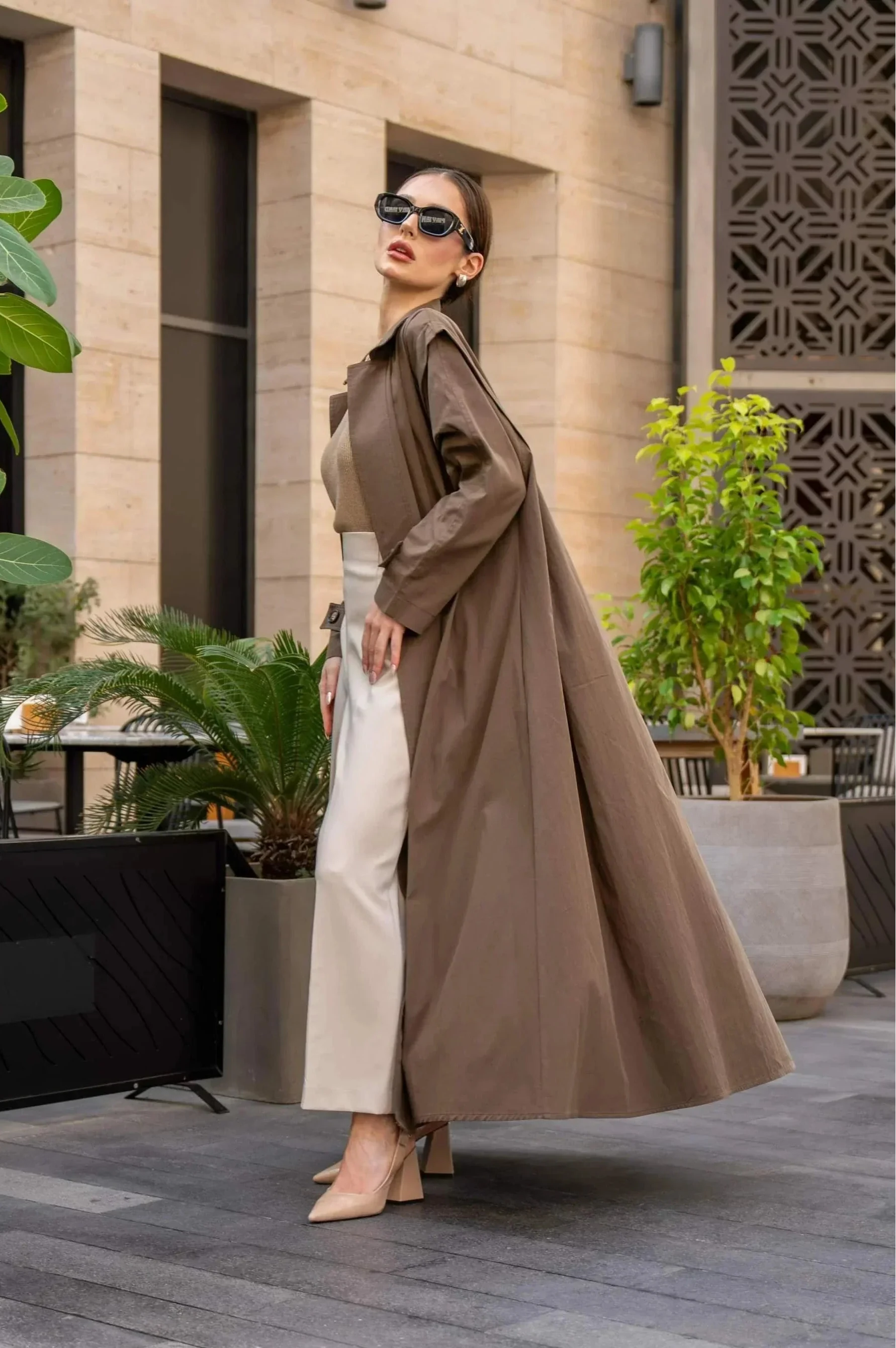 Woman wearing a long coffee chino coat with tailored fit and sunglasses in an outdoor urban setting