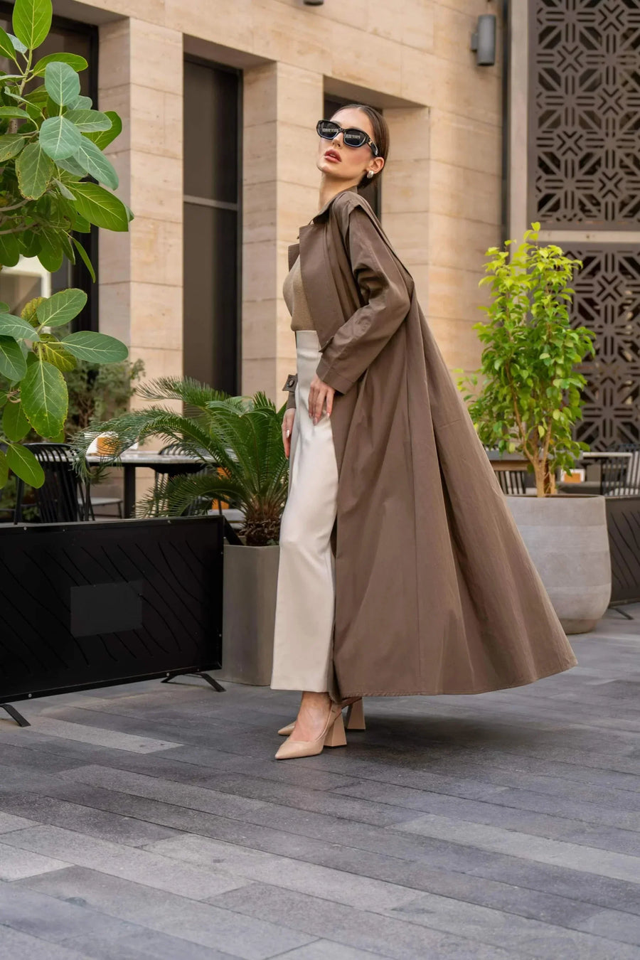Woman wearing a long coffee chino coat with tailored fit and sunglasses in an outdoor urban setting