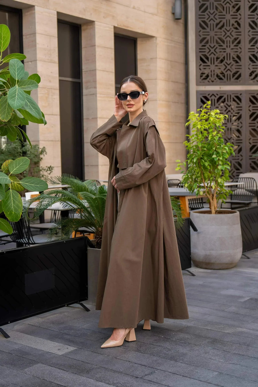 Woman wearing a long coffee chino coat with tailored fit and sunglasses in an outdoor urban setting