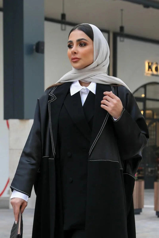 Woman wearing a black abaya made of premium mokado fabric with white contrasting stitching and a light-colored hijab outdoors.