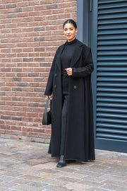 Woman wearing a black crepe abaya with a long black coat standing outdoors against a brick wall and blue door