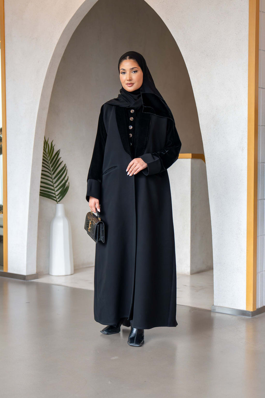 Elegant black refined abaya with velvet details, gold buttons, and blazer-style lapel, worn by a woman in a modern archway setting