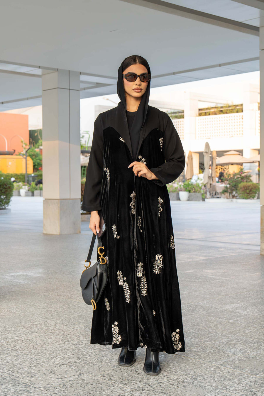 Black Velvet Bloom abaya with intricate embroidery, loose bisht-style cut, and hood, worn with black boots and sunglasses outdoors.