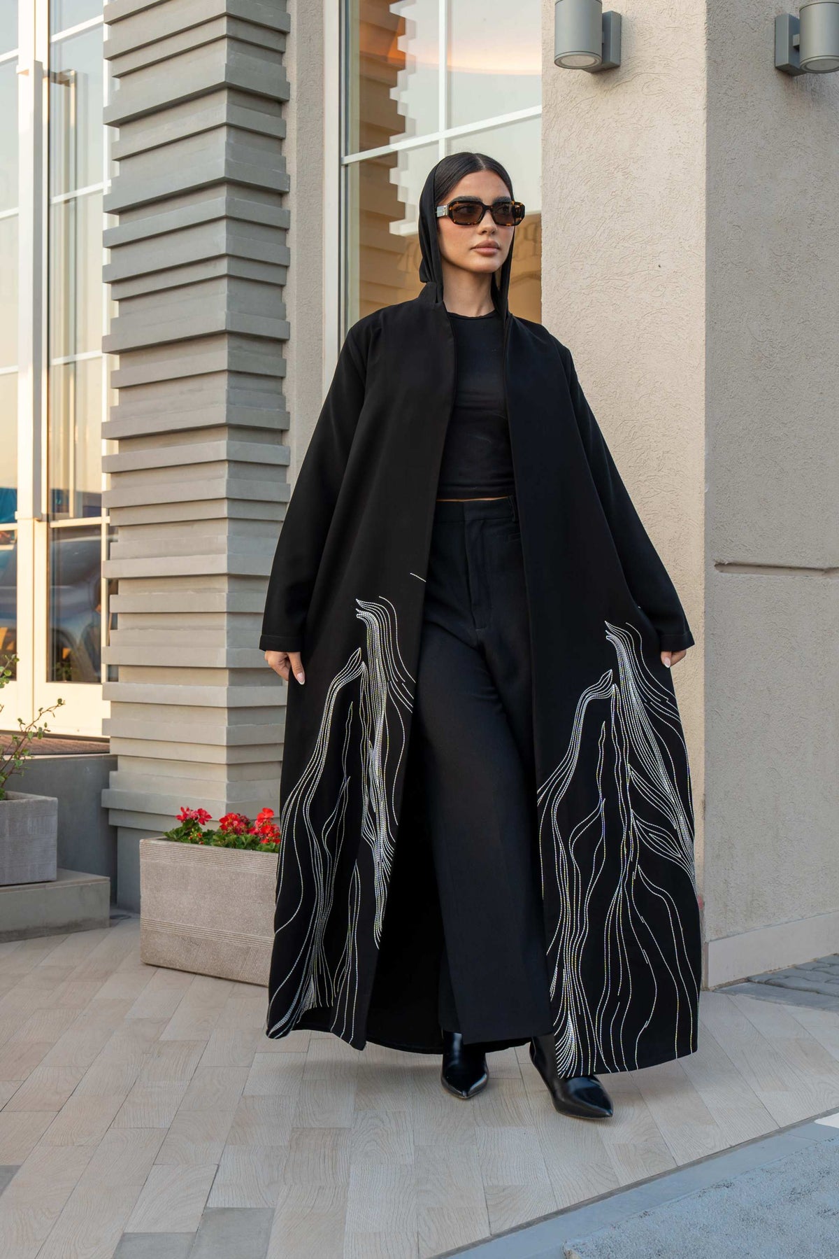 Woman wearing a black crepe abaya with white shak embroidery along the lower edges, standing outdoors near modern architecture