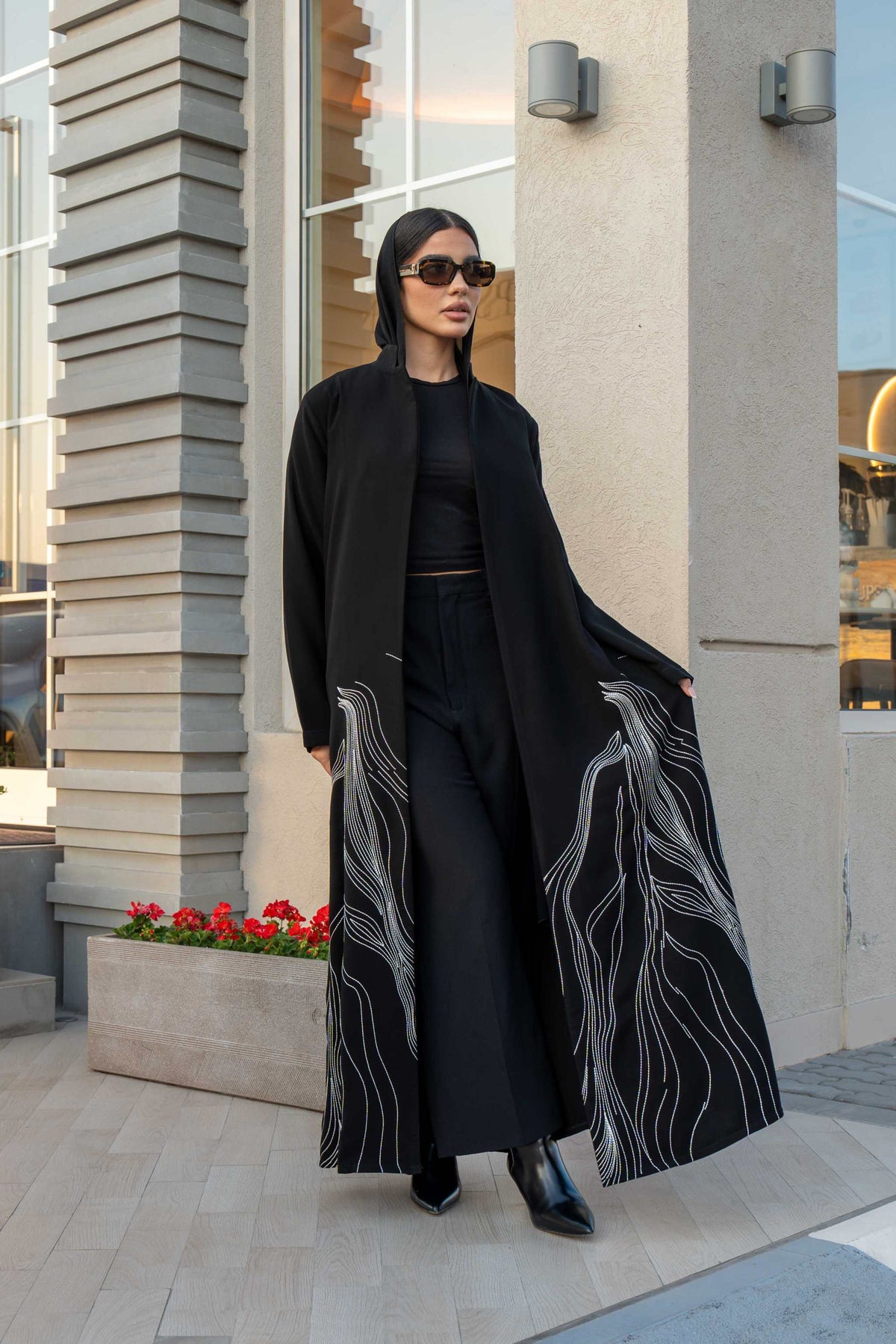 Woman wearing a black crepe abaya with white shak embroidery along the lower edges, standing outdoors near modern architecture