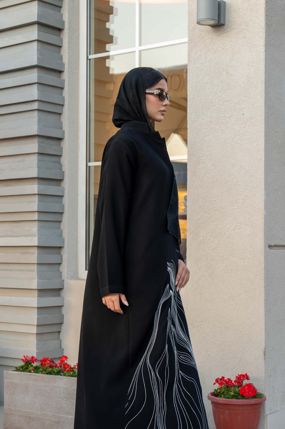 Woman wearing a black crepe abaya with white shak embroidery along the lower edges, standing outdoors near modern architecture