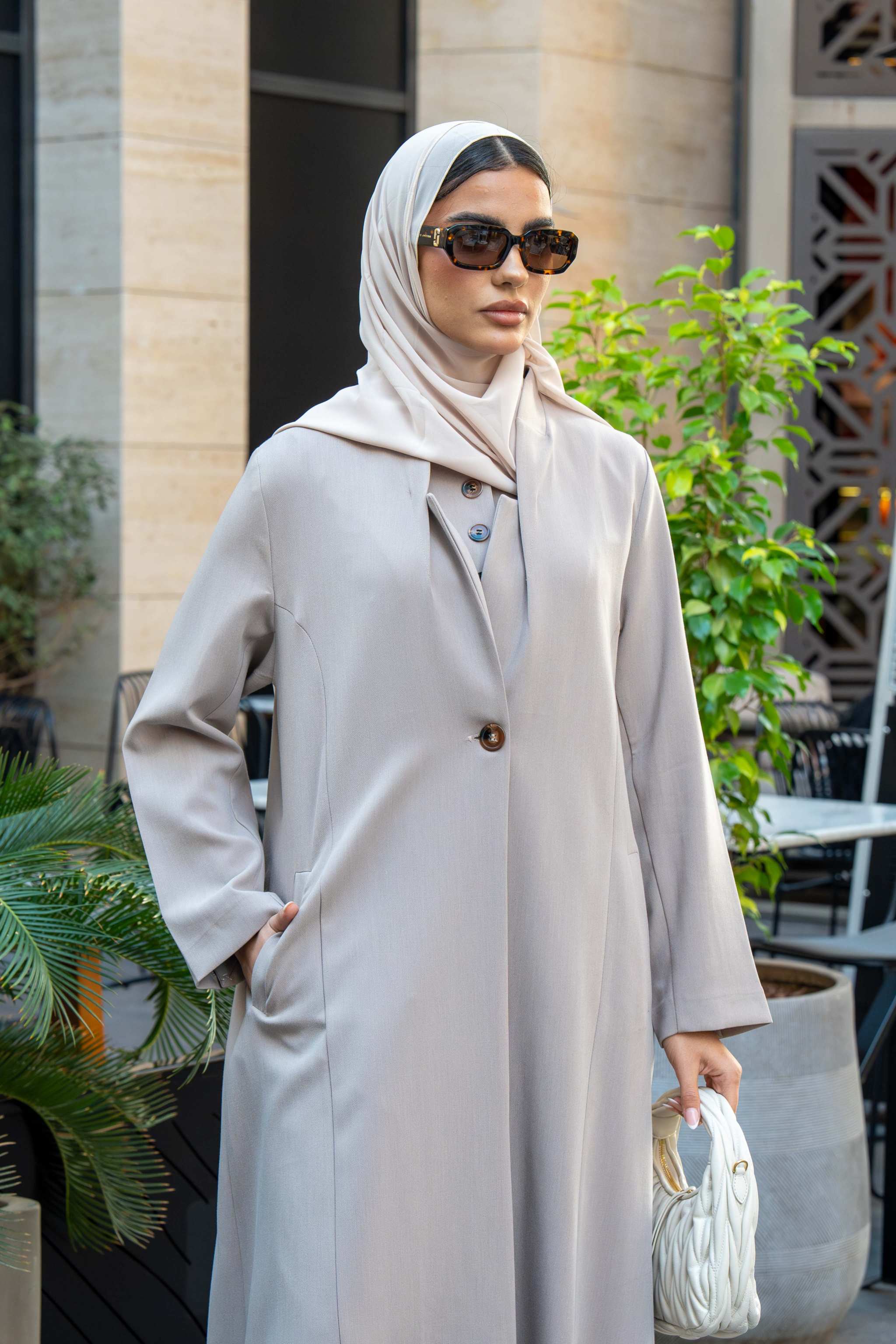 Woman wearing a beige crepe abaya with a single front button and flowy silhouette holding a white handbag outdoors