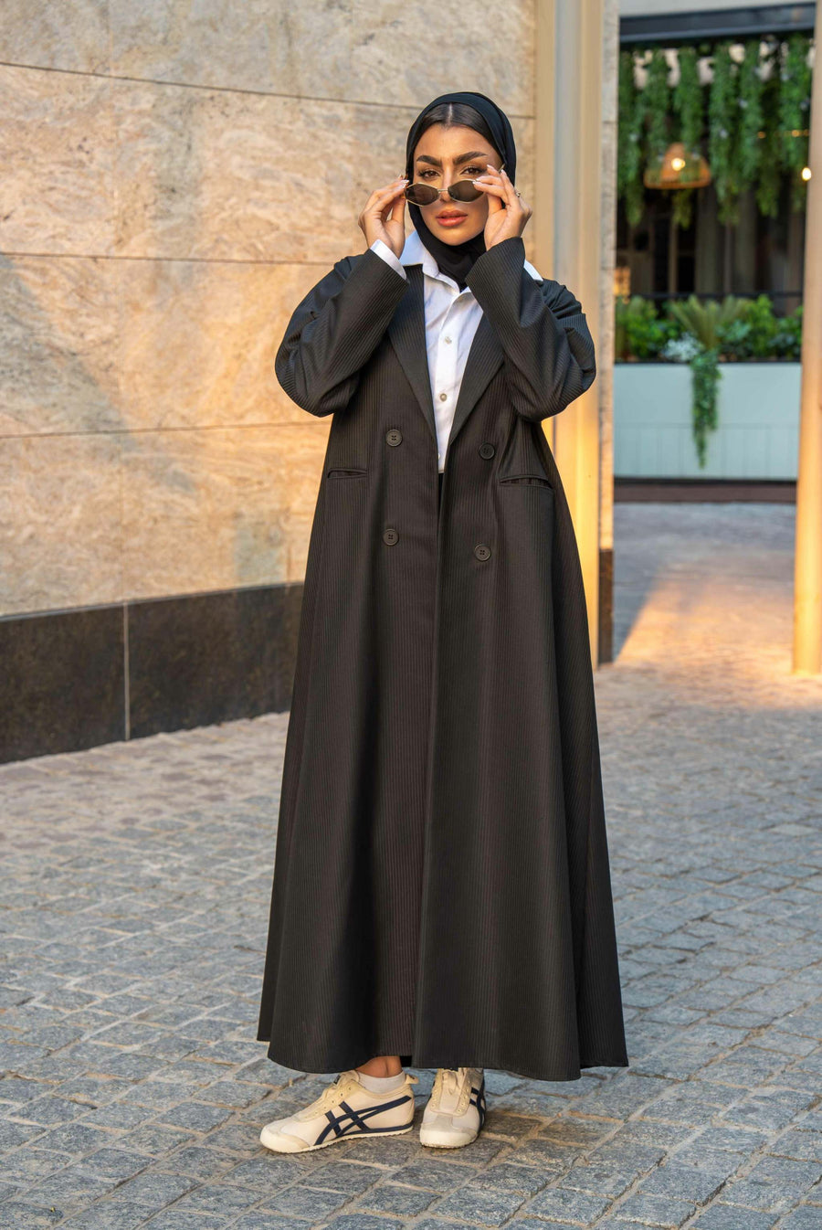 Woman wearing a structured black crepe abaya with pinstripe texture and double-breasted front, styled with sneakers and sunglasses