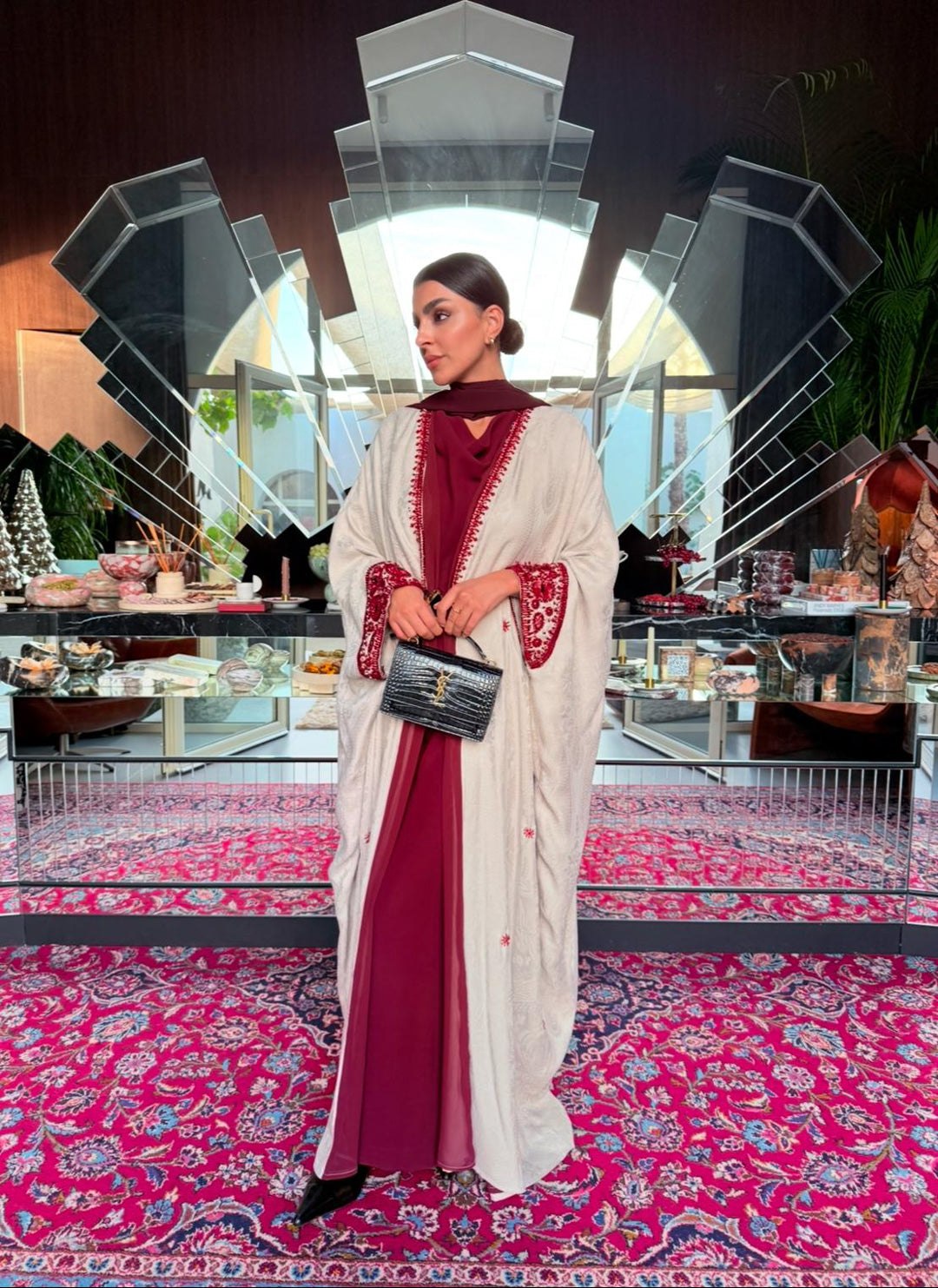 Woman wearing Harir Kubra Ivory Abaya with burgundy embroidery on sleeves and front edges in elegant indoor setting
